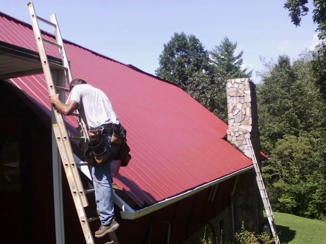 Roofer on a ladder beside a red metal roof and stone chimney — Soffit & Fascia Repair by Roofing Brothers in Cadogan